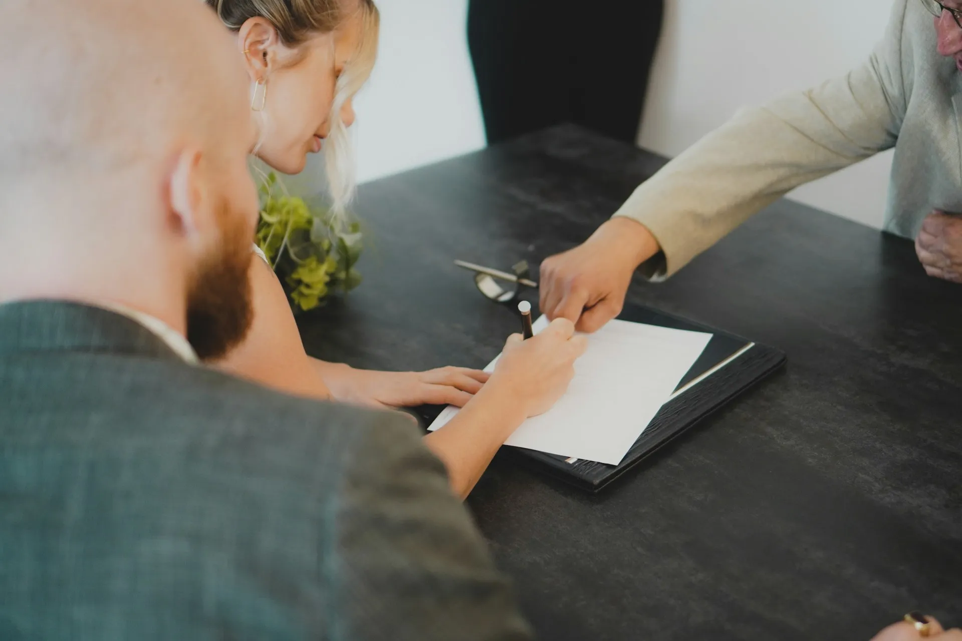 A couple signing a legal document at a desk, assisted by a third party