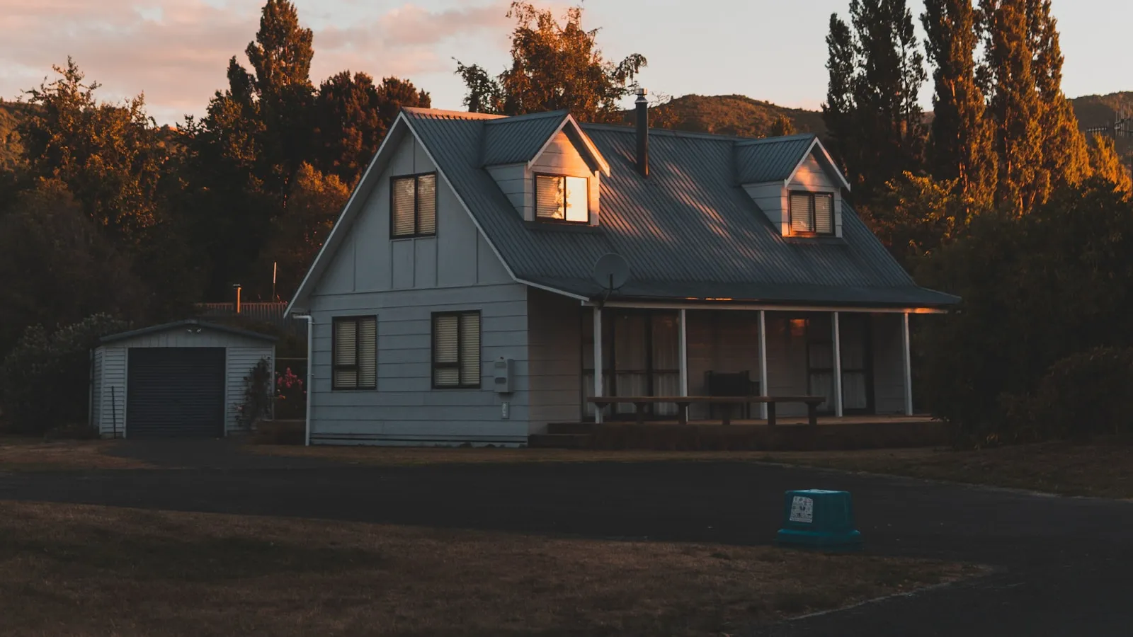 A New Zealand weatherboard house with lit windows at dusk, set back from the road with a garage and driveway